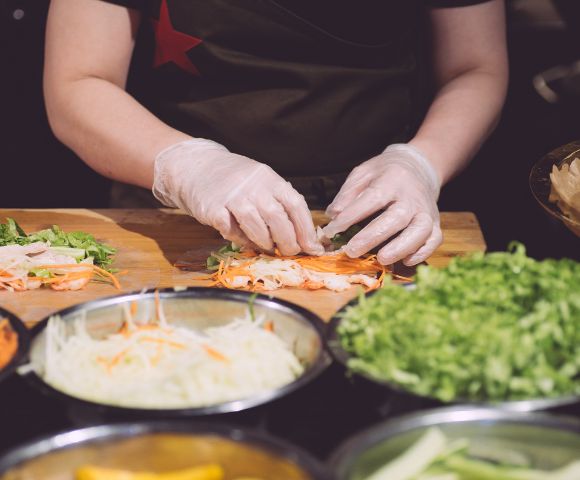 A chef in gloves prepares fresh spring rolls, carefully layering ingredients like shrimp, lettuce, and carrots on a wooden counter.