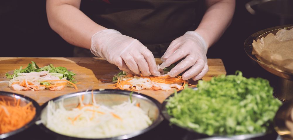 A chef in gloves prepares fresh spring rolls, carefully layering ingredients like shrimp, lettuce, and carrots on a wooden counter.