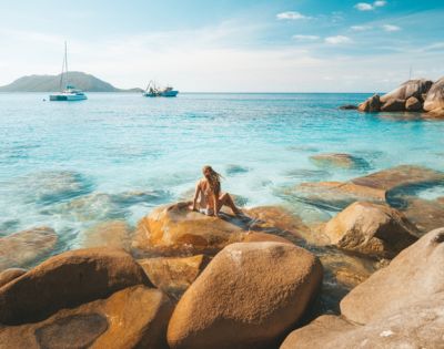 Person sitting on rocks by the clear blue sea with another person swimming nearby.