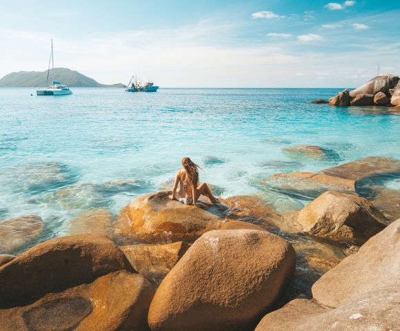 Person sitting on rocks by the clear blue sea with another person swimming nearby.