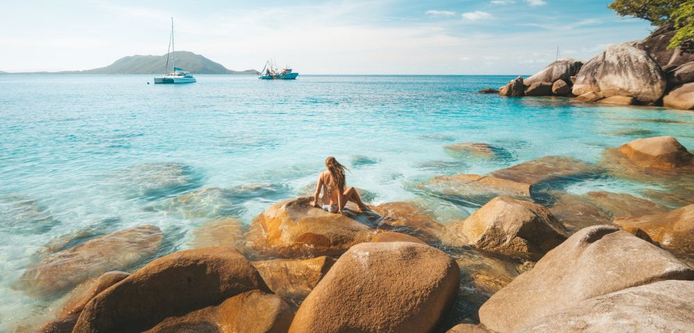 Person sitting on rocks by the clear blue sea with another person swimming nearby.