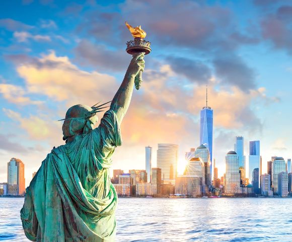 Statue of Liberty in the foreground with a raised torch, overlooking the New York City skyline across calm water during a vibrant sunset. Skyscrapers are bathed in warm light under a partly cloudy sky.