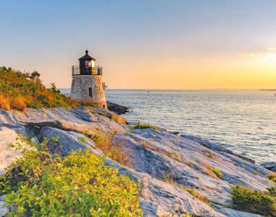 A stone lighthouse on a rocky cliff at sunset, overlooking a calm ocean. The sky is golden, and lush greenery surrounds the foreground rocks.