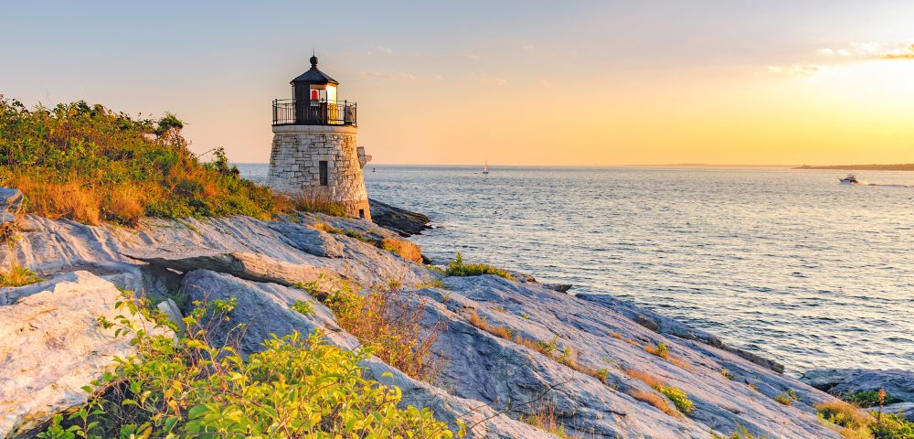 A stone lighthouse on a rocky cliff at sunset, overlooking a calm ocean. The sky is golden, and lush greenery surrounds the foreground rocks.