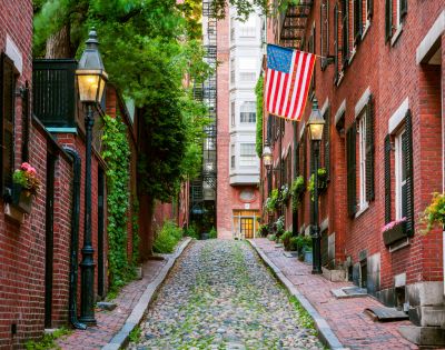 Narrow cobblestone street between red brick buildings adorned with lush greenery, vintage lamps, and a large American flag, creating a charming, historical ambiance.