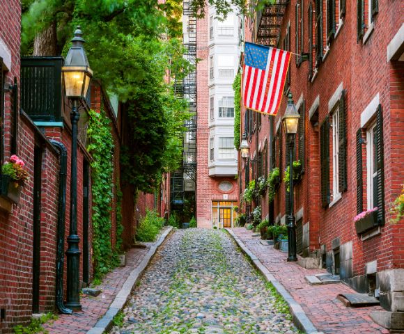 Narrow cobblestone street between red brick buildings adorned with lush greenery, vintage lamps, and a large American flag, creating a charming, historical ambiance.