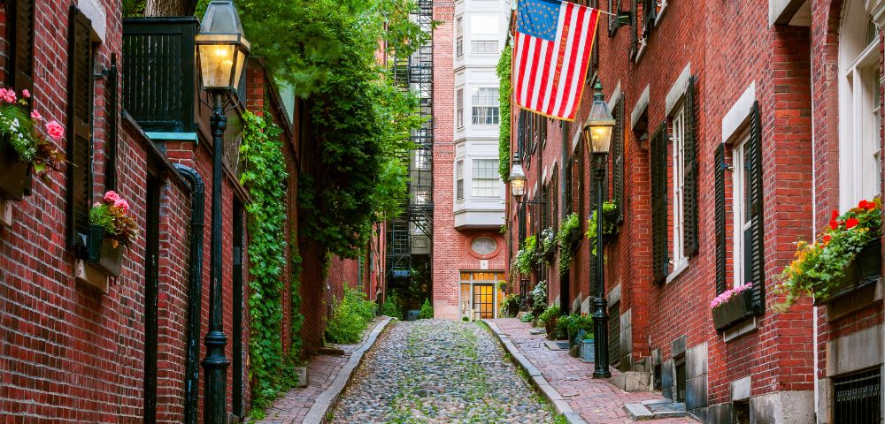 Narrow cobblestone street between red brick buildings adorned with lush greenery, vintage lamps, and a large American flag, creating a charming, historical ambiance.