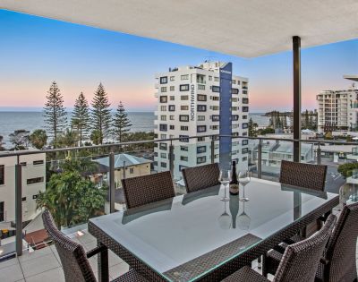 An outdoor dining area on a balcony overlooking buildings, trees, and the ocean during a sunset.