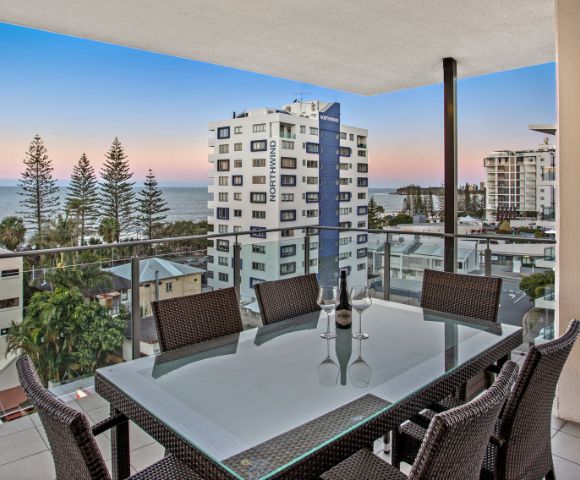 An outdoor dining area on a balcony overlooking buildings, trees, and the ocean during a sunset.
