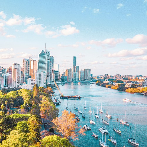 Aerial view of a vibrant city skyline with tall skyscrapers, lush green parks, and a wide river dotted with boats under a bright, partly cloudy sky.