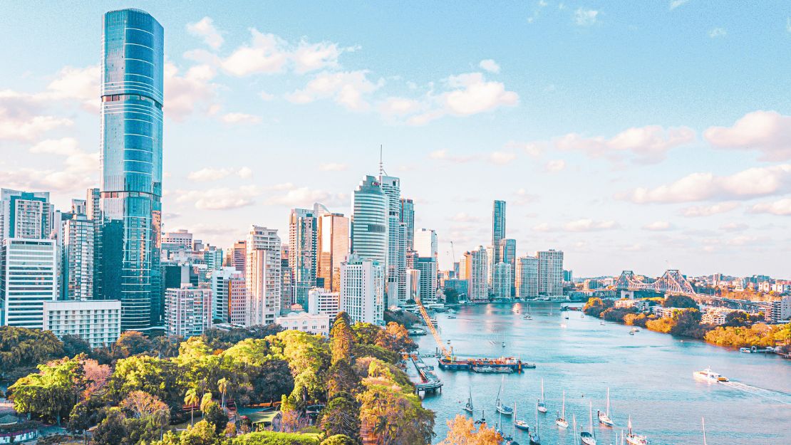 Aerial view of a vibrant city skyline with tall skyscrapers, lush green parks, and a wide river dotted with boats under a bright, partly cloudy sky.