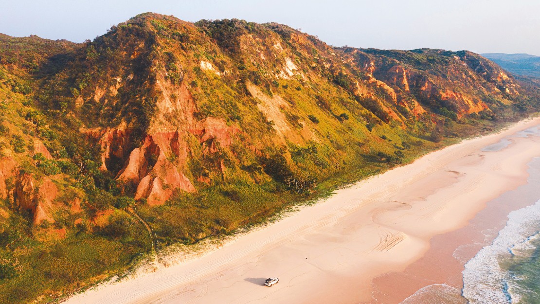 Aerial view of a vast, empty beach bordered by towering, sunlit red cliffs with lush greenery. A lone vehicle drives on the sandy shore, evoking solitude.