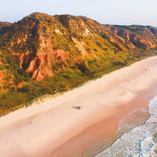 Aerial view of a vast, empty beach bordered by towering, sunlit red cliffs with lush greenery. A lone vehicle drives on the sandy shore, evoking solitude.