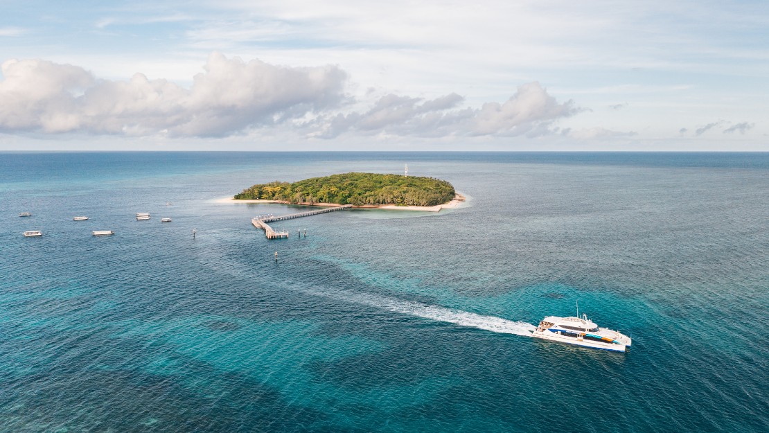 Aerial view of a boat cruising on clear blue waters away from a small, lush green island with a pier.