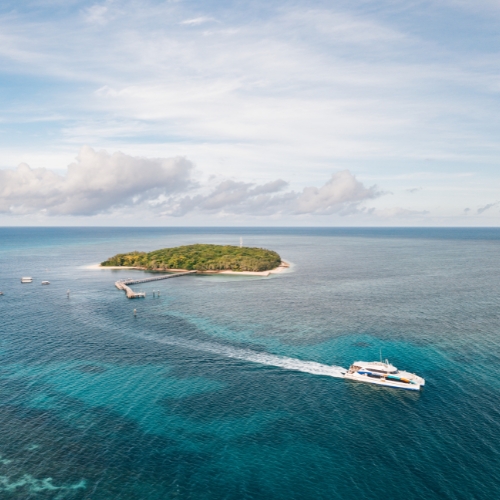Aerial view of a boat cruising on clear blue waters away from a small, lush green island with a pier.