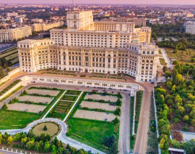 Aerial view of a large, ornate palace surrounded by manicured gardens and tree-lined paths under a clear sky, with a sprawling cityscape in the background.