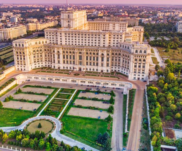 Aerial view of a large, ornate palace surrounded by manicured gardens and tree-lined paths under a clear sky, with a sprawling cityscape in the background.