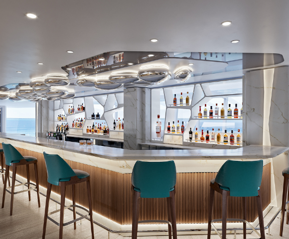 A modern bar with teal stools, elegant lighting, and shelves lined with various bottles, featuring a coastal view in the background.