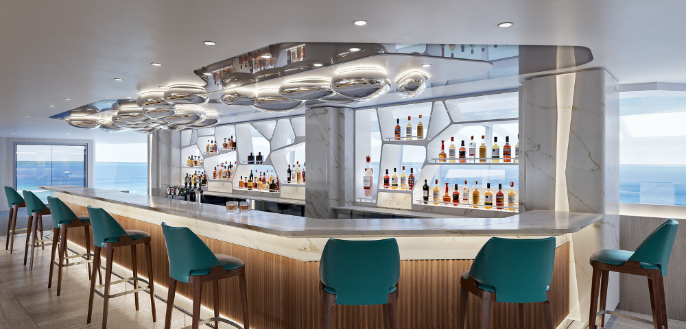 A modern bar with teal stools, elegant lighting, and shelves lined with various bottles, featuring a coastal view in the background.