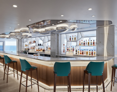 A modern bar with teal stools, elegant lighting, and shelves lined with various bottles, featuring a coastal view in the background.