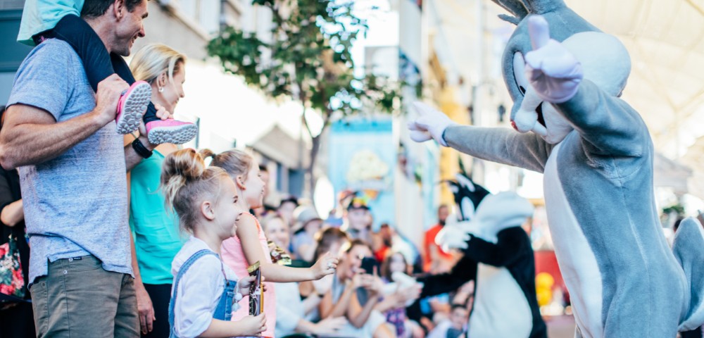 Children excitedly greet a person in a Bugs Bunny costume during a lively outdoor event, surrounded by a cheerful crowd and colorful decor.