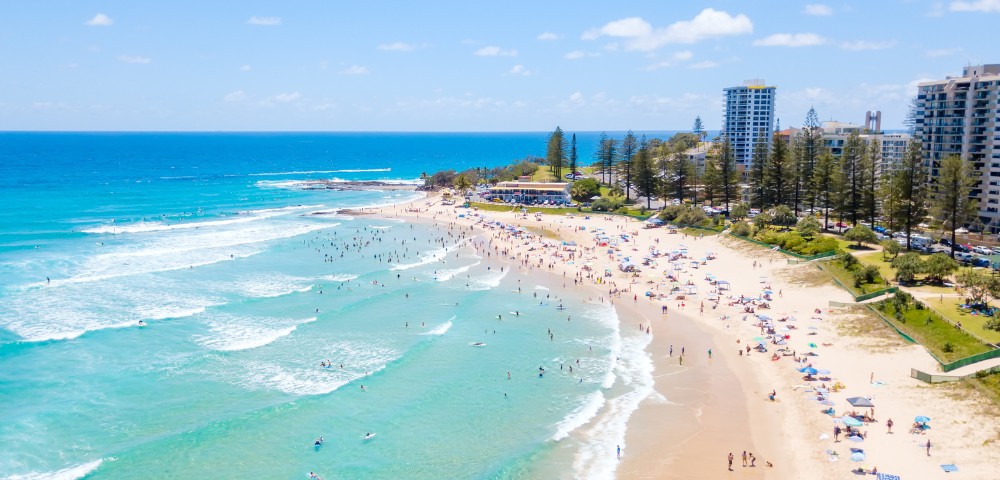 Aerial view of a vibrant beach scene with turquoise waves, sunbathers, and surfers. High-rise buildings and green trees line the background under a clear blue sky.