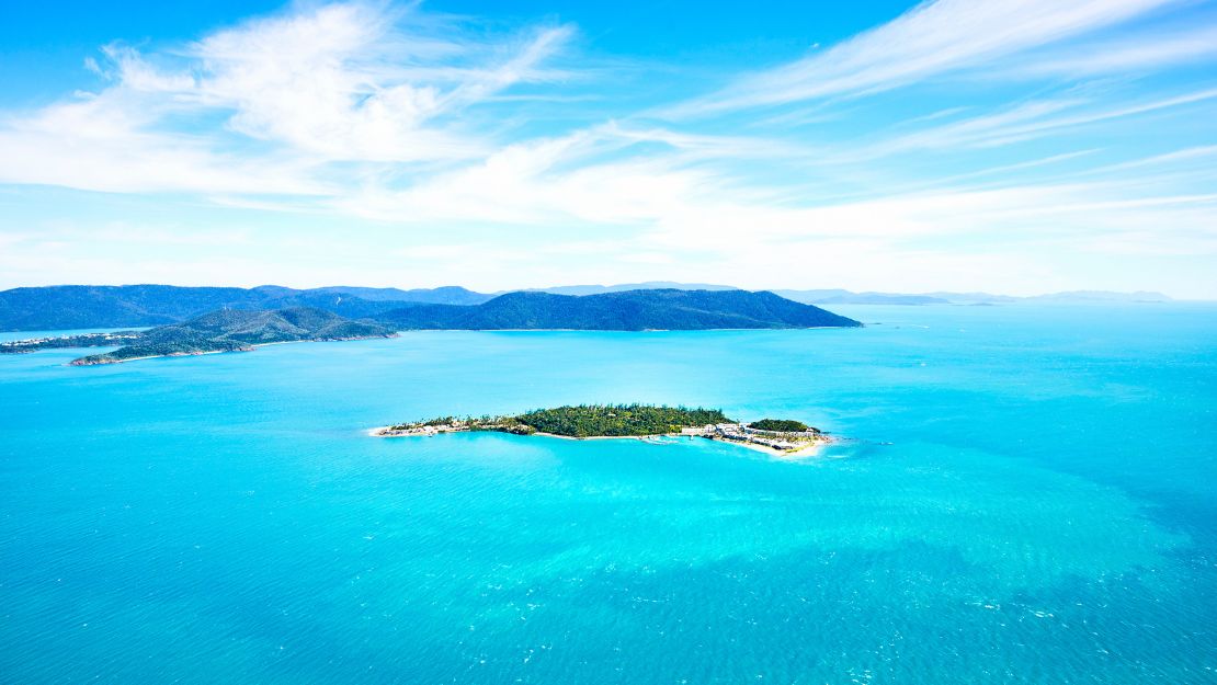 A small, lush green island surrounded by vibrant turquoise waters sits under a clear blue sky with wispy clouds.