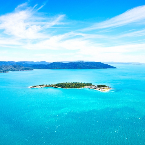 A small, lush green island surrounded by vibrant turquoise waters sits under a clear blue sky with wispy clouds.