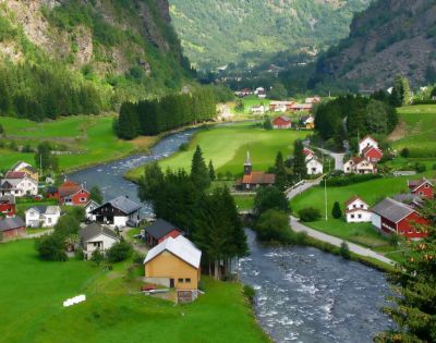 Scenic view of a quaint village with white and red houses nestled in lush green valleys. A clear river winds through the landscape, with forested hills in the background.