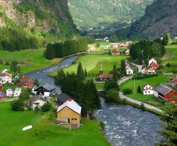 Scenic view of a quaint village with white and red houses nestled in lush green valleys. A clear river winds through the landscape, with forested hills in the background.