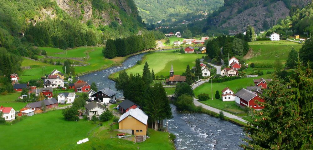 Scenic view of a quaint village with white and red houses nestled in lush green valleys. A clear river winds through the landscape, with forested hills in the background.