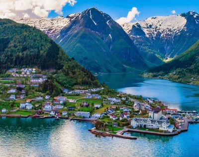 Scenic view of a fjord with turquoise water surrounded by snow-capped mountains. A charming village with colorful houses lines the verdant shoreline.