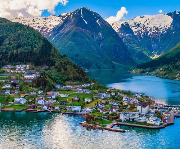 Scenic view of a fjord with turquoise water surrounded by snow-capped mountains. A charming village with colorful houses lines the verdant shoreline.
