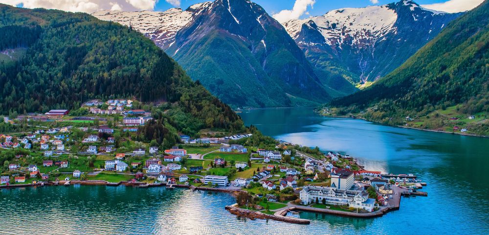 Scenic view of a fjord with turquoise water surrounded by snow-capped mountains. A charming village with colorful houses lines the verdant shoreline.