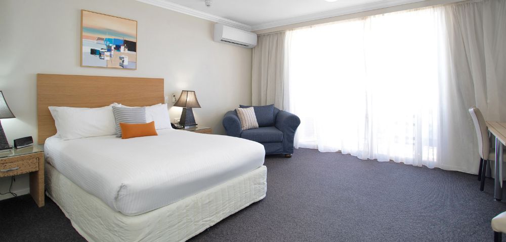 Hotel Room Interior – A simple hotel room with a bed, bedside table, lamp, and a large curtained window.