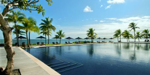 A tranquil resort pool with palm trees, sun loungers, and umbrellas, overlooking calm blue waters and a clear sky.