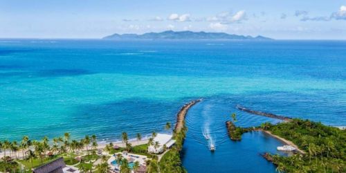 Aerial view of a tropical coastline with turquoise waters, palm trees, and a river leading to the ocean, set against distant islands.