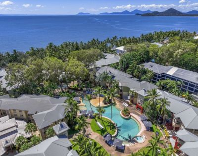 Aerial view of a tropical resort with a pool surrounded by lush greenery and the ocean in the background.