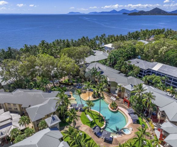 Aerial view of a tropical resort with a pool surrounded by lush greenery and the ocean in the background.