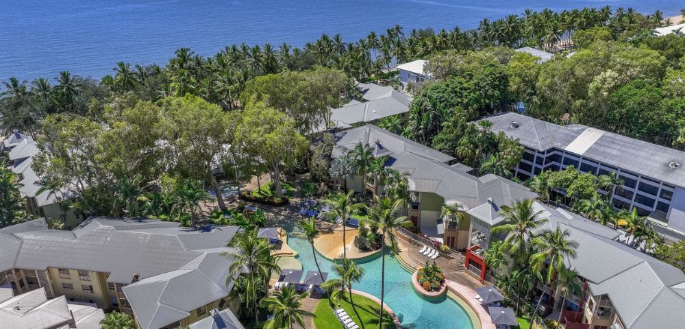 Aerial view of a tropical resort with a pool surrounded by lush greenery and the ocean in the background.