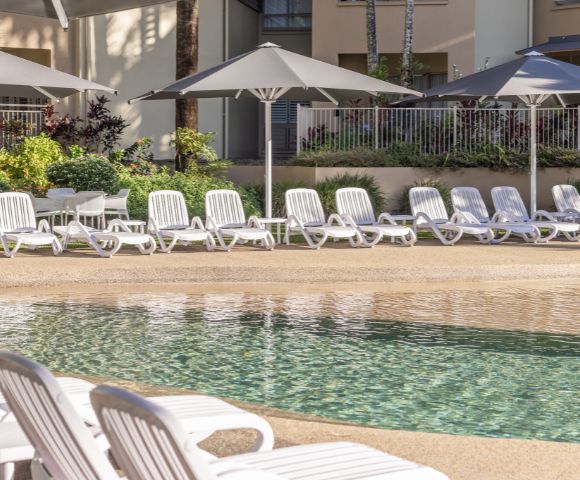 Row of white lounge chairs next to a swimming pool with umbrellas in the background.