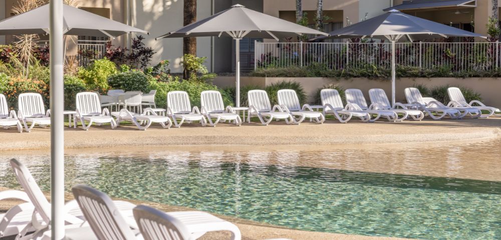 Row of white lounge chairs next to a swimming pool with umbrellas in the background.