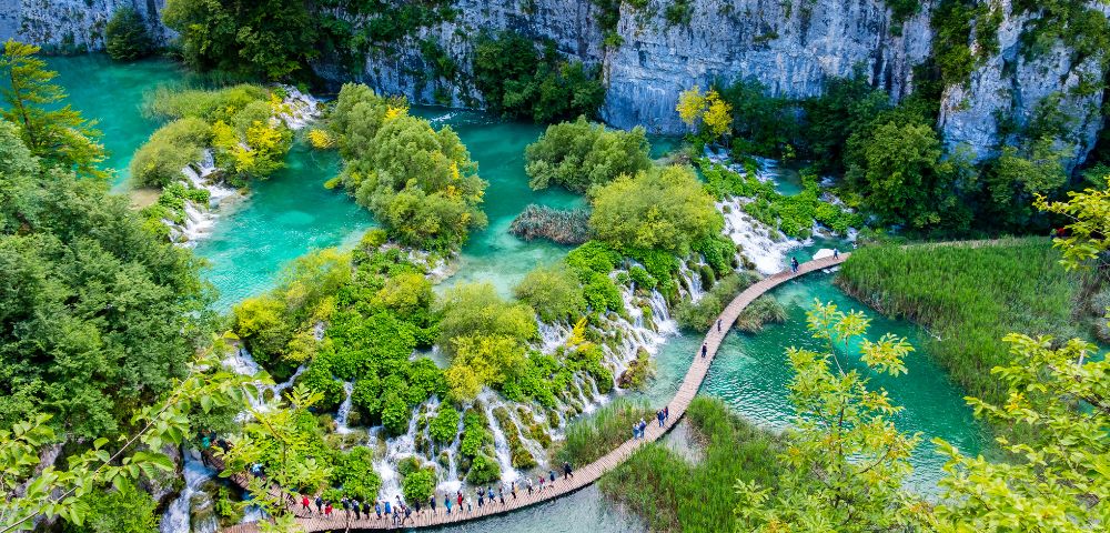 Vibrant aerial view of Plitvice Lakes, Croatia, featuring turquoise waters, lush green foliage, and a winding wooden boardwalk with visitors exploring.