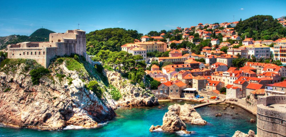 Coastal cityscape with a historic fortress atop a rocky cliff, overlooking turquoise waters. Nearby, houses with red-tiled roofs under a clear blue sky.