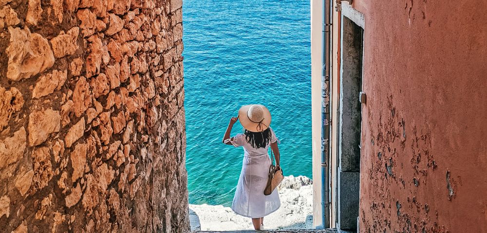 A person in a white dress and sun hat descends stone steps between rustic walls toward a bright blue sea, evoking a sense of serene adventure.