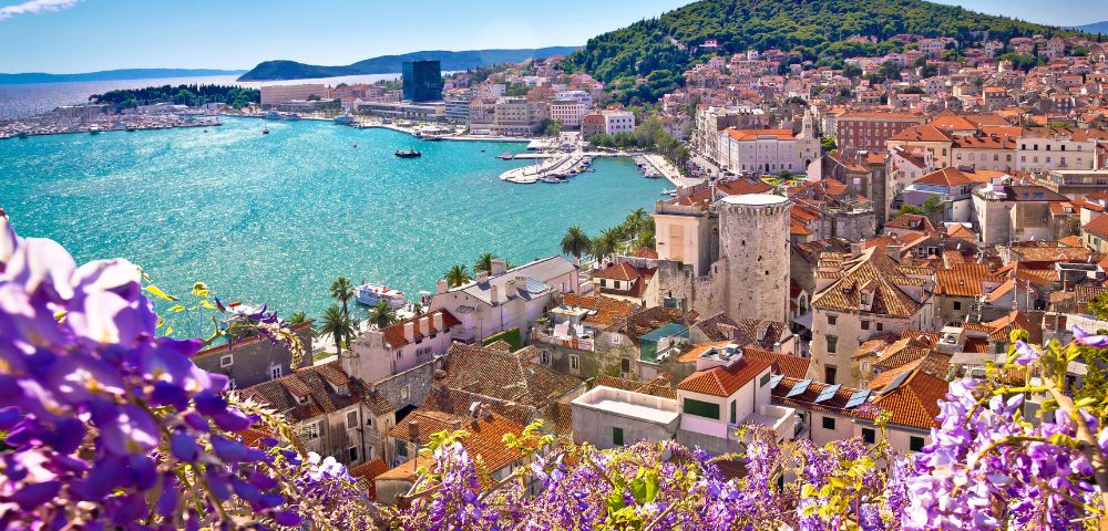 Scenic view of a coastal town with terracotta rooftops and historic stone buildings. The turquoise sea sparkles under the sun, surrounded by green hills and purple flowers in the foreground.