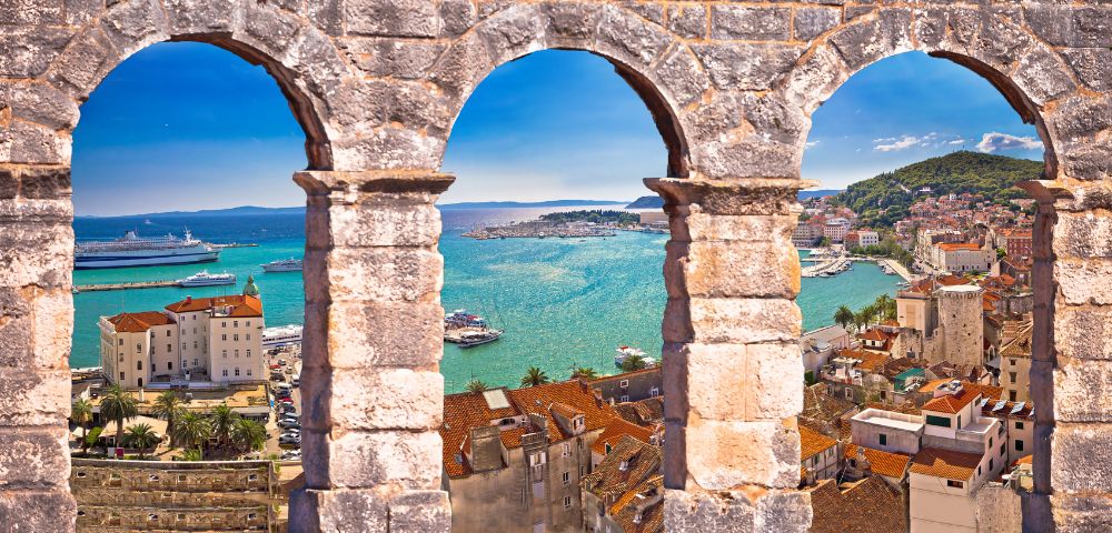 View of a coastal town from a stone wall with three arches. Turquoise sea, boats, and red-tiled roofs under a vibrant blue sky create a serene scene.