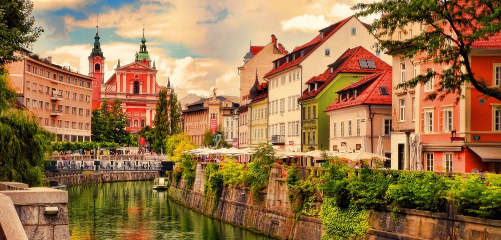 Colorful riverside buildings line a peaceful canal in Ljubljana. The sky is partly cloudy, and a prominent pink church stands in the background.