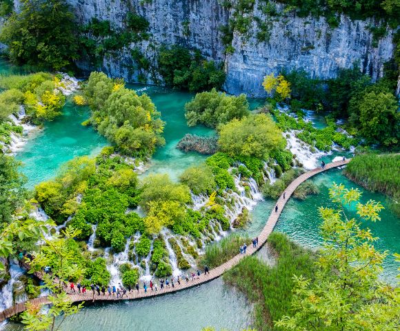 Vibrant aerial view of Plitvice Lakes, Croatia, featuring turquoise waters, lush green foliage, and a winding wooden boardwalk with visitors exploring.