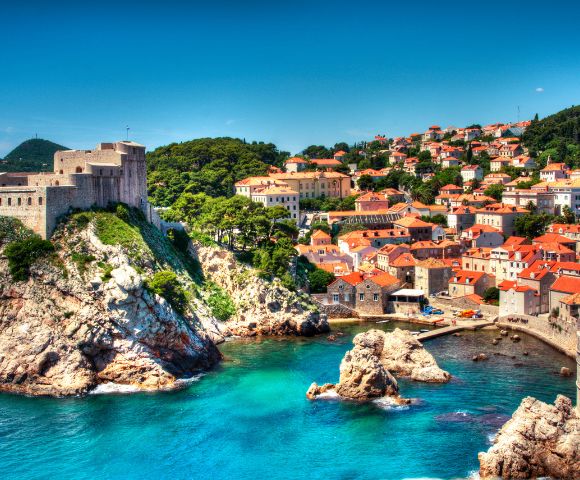 Coastal cityscape with a historic fortress atop a rocky cliff, overlooking turquoise waters. Nearby, houses with red-tiled roofs under a clear blue sky.